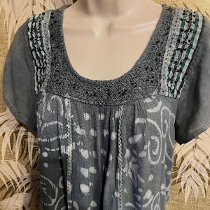 May include: Gray short-sleeve top with a scoop neckline. The top features a white and gray patterned design on the front and a decorative neckline with black sequins and green accents. The sleeves are short and the fabric appears to be lightweight.