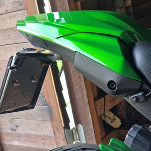 May include: A bright green motorcycle tail fairing with a black license plate holder. The fairing has a sleek, aerodynamic design and is mounted on a black motorcycle seat. The background is a wooden structure.
