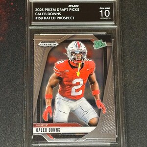 May include: A graded Caleb Downs football card from the 2025 Prizm Draft Picks series. The card features Downs in an Ohio State Buckeyes uniform, graded GEM MINT 10. The card is encased in a protective holder.