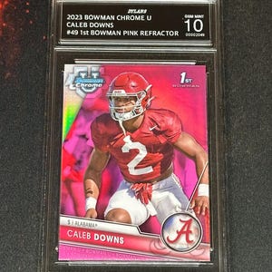 May include: A Caleb Downs 2023 Bowman Chrome U football card, graded Gem Mint 10. The card features a pink refractor design and the text "#49 1st Bowman Pink Refractor". The player is wearing a red uniform with the number 2.