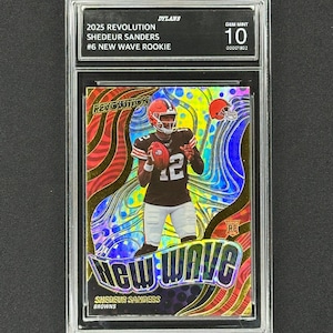 May include: A graded trading card featuring Shedeur Sanders in a Browns uniform. The card is from the 2025 Revolution series, labeled as a New Wave Rookie. The card is graded Gem Mint 10 and encased in a protective holder.