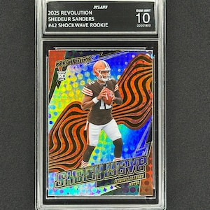 May include: A professionally graded trading card of Shedeur Sanders in a brown and white uniform. The card is labelled "2025 Revolution" and "Shockwave Rookie." The card is graded Gem Mint 10 and encased in a protective holder.