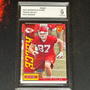 May include: A graded sports trading card featuring Travis Kelce in a red Kansas City Chiefs uniform. The card is from the 2013 Rookies & Stars series, with the text "#192 ROOKIE" visible. The card is graded MINT 9.