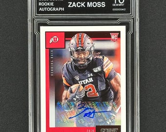 May include: A graded trading card of Zack Moss, a running back for the Utah Utes. The card is a 2020 Score Rookie Autograph card, graded a 10 by BGS. The card features a photo of Moss in a Utah uniform, running with the football. The card has a red, white, and black colour scheme. The card is signed by Moss.