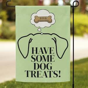 Pode incluir: Uma bandeira de jardim verde claro com o texto "HAVE SOME DOG TREATS!" em preto. Acima do texto está o contorno da cabeça de um cão e um balão de pensamento com um petisco em forma de osso. A bandeira é exibida em um suporte de metal preto.