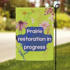 Puede incluir: Una bandera de jardín verde con un diseño de acuarela de flores y el texto "Prairie restoration in progress".