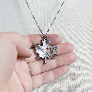 May include: A silver necklace with a detailed maple leaf pendant. The leaf has a heart-shaped cutout in the center.