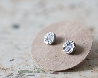 May include: Two silver textured round stud earrings displayed on a brown paper circle.