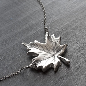 Puede incluir: Un collar con un colgante de hoja de arce plateado y una cadena delicada. La hoja está detallada con venas y una superficie texturizada.