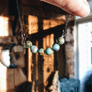 May include: A turquoise and green stone necklace with a silver chain. The necklace has seven stones, each with a different pattern.