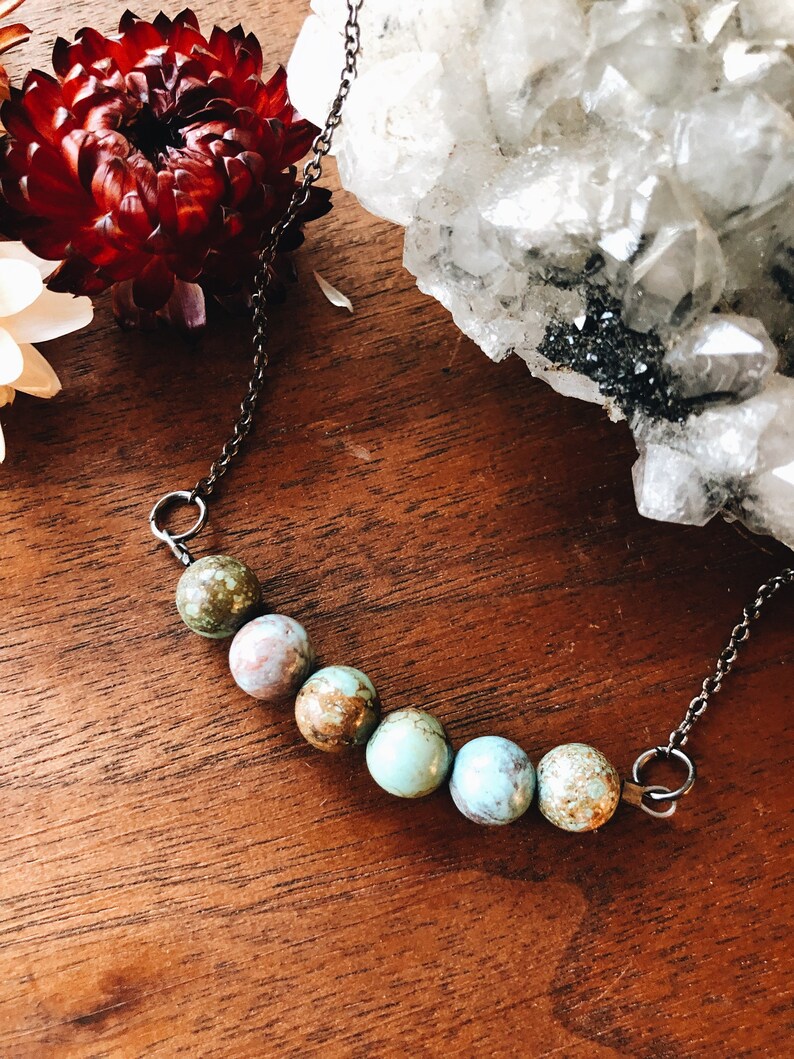 May include: A silver chain necklace with six turquoise and brown gemstone beads. The necklace is on a wooden surface.