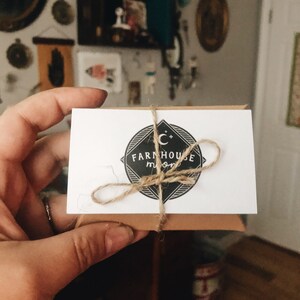 K&ouml;nnte beinhalten: Eine wei&szlig;e Karte mit einer braunen Kordelschleife darum gebunden. Die Karte hat ein schwarz-wei&szlig;es Logo mit dem Text "FARMHOUSE MOON" und einen Halbmond mit Sternen.