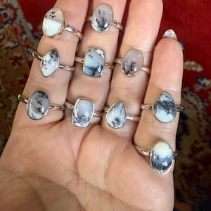 May include: A collection of silver rings, each featuring a unique oval or teardrop-shaped stone with black and white dendritic patterns. The rings are displayed on a hand, showcasing the variety of designs and sizes.