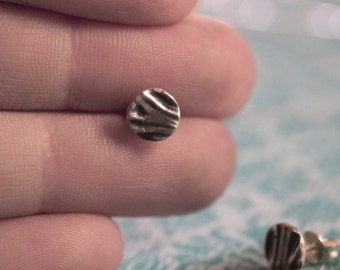 May include: A pair of silver stud earrings with a textured, circular design. The earrings are shown on a person's finger.