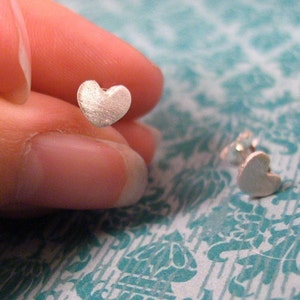 May include: A pair of silver heart-shaped stud earrings. The earrings are small and delicate, and they have a slightly textured surface.