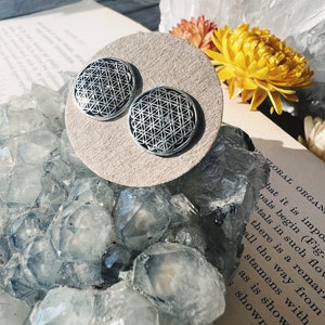Puede incluir: Un par de pendientes de botón plateados con un diseño geométrico de la flor de la vida. Los pendientes se exhiben sobre una superficie circular beige, con un fondo de cristales, flores y un libro antiguo.
