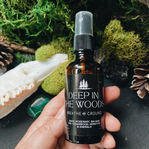 May include: A brown glass bottle with a black spray nozzle and a white label that reads "DEEP IN THE WOODS BREATHE & GROUND SAGE, ROSEMARY, BALSAM FIR, CEDARWOOD, HEMATITE & EMERALD"