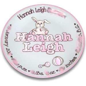 May include: A white ceramic baby plate with pink trim and a pink and white striped design. The plate features a bunny holding a heart, a rattle, a ball, and the text "Hannah Leigh".