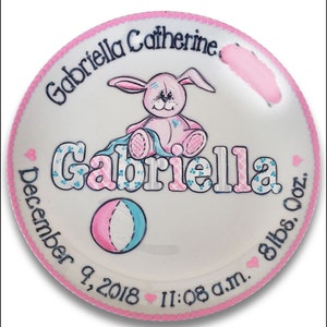 May include: A white ceramic plate with a pink border and a pink bunny holding a blue and pink blanket. The plate is decorated with the name "Gabriella Catherine" and the date "December 9, 2018" along with the time "11:08 a.m." and the weight "8 lbs. 0 oz."