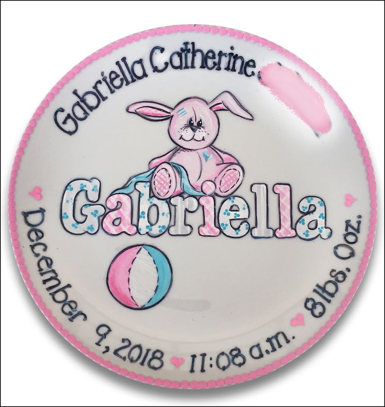 May include: A white ceramic plate with a pink border and a pink bunny holding a blue and pink blanket. The plate is decorated with the name "Gabriella Catherine" and the date "December 9, 2018" along with the time "11:08 a.m." and the weight "8 lbs. 0 oz."