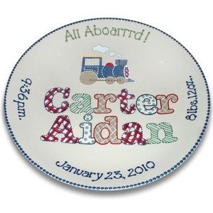May include: A white ceramic plate with blue trim and a train design. The plate is personalized with the names "Carter Aidan", the date "January 23, 2010", the time "6:56pm", and the weight "8lbs 12oz".