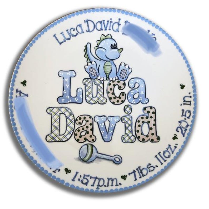 May include: A white ceramic plate with a blue border and a blue dinosaur design. The plate is personalized with the name "Luca David" and the birth details: 1:57 p.m., 7 lbs 11 oz, 20.5 in.