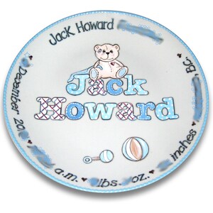 May include: A white ceramic plate with a blue border, decorated with a teddy bear, a rattle, and a ball. The plate is personalized with the name "Jack Howard" and the date "December 20".
