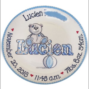 May include: A white ceramic plate with a blue border and a blue and white checkered design. The plate features a teddy bear, a ball, and the name "Lucien" written in blue and white. The plate also includes the date "November 20, 2018", the time "11:48 a.m.", and the baby's weight and length: "7 lbs. 8 oz. 54 cm."