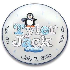 May include: A white ceramic plate with a blue border, featuring a hand-painted penguin with a scarf, the names "Tyler Jack", the date "July 7, 2016", and the birth details "7 lbs. 19 in. 7:39 am."