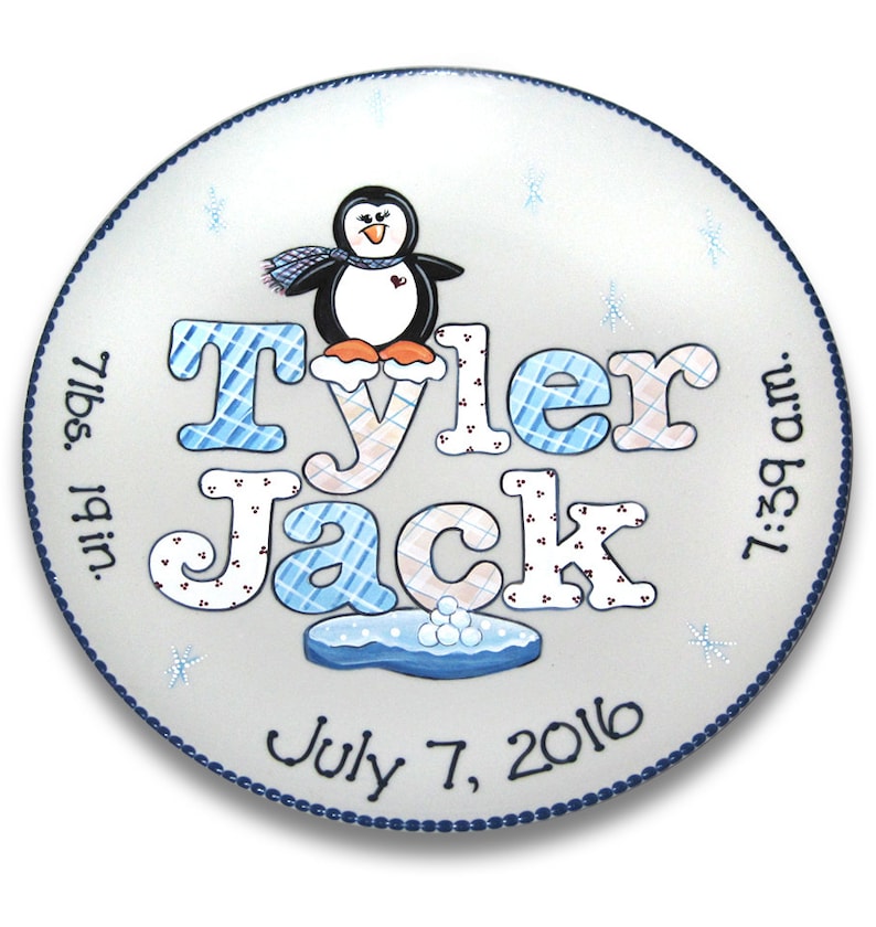 May include: A white ceramic plate with a blue border, featuring a hand-painted penguin with a scarf, the names "Tyler Jack", the date "July 7, 2016", and the birth details "7 lbs. 19 in. 7:39 am."