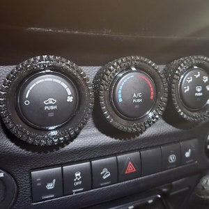 May include: Black car dashboard with three circular control knobs. The knobs have textured, decorative rings around them. The center knob is labeled "A/C" and "PUSH". Other buttons include "PUSH" and icons for fan and air direction.