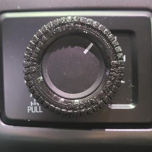 May include: Close-up of a black textured knob with a white indicator line, set against a dark dashboard. The knob is surrounded by a textured ring. The word "PULL" is visible below the knob.