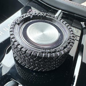 May include: A circular black knob cover with a textured tire tread pattern on the outer edge and a smooth, brushed silver metallic center disc. The object is designed to fit over a vehicle's existing control dial, featuring a rugged, rubberized appearance.