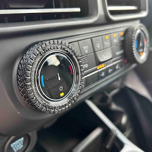 May include: Black textured tire-shaped covers for car climate control knobs. The covers are installed on a car's dashboard, surrounding the knobs. The dashboard has buttons and a digital display. The car's interior is black.