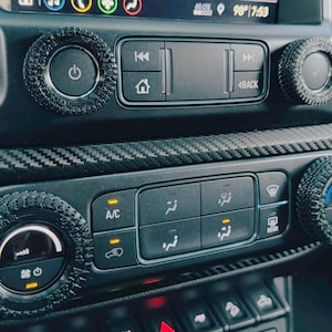 May include: Close-up of a car's dashboard with black and silver controls. The controls include a power button, a home button, a back button, and a temperature control knob. The dashboard is made of carbon fiber.