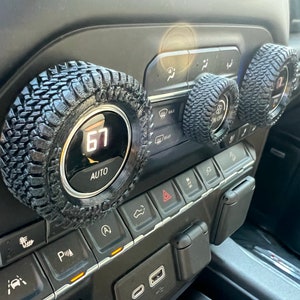 May include: Close-up of a car's dashboard with a digital display showing "67" and the word "AUTO". The display is surrounded by a black tire-tread-patterned knob. The dashboard also features other knobs and buttons.