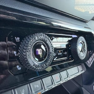 May include: Close-up of a car's dashboard with two round knobs that have a tire tread design. The knobs are black and have a silver ring around the edge. The knob on the left has a digital display that reads "70".