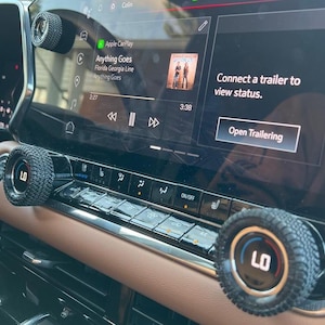 May include: A car's dashboard with a touchscreen display showing the song "Anything Goes" by Florida Georgia Line playing on Apple CarPlay. The screen also shows a message that says "Connect a trailer to view status." and a button that says "Open Trailering". There are three black and silver knobs with a tire tread design on the dashboard.