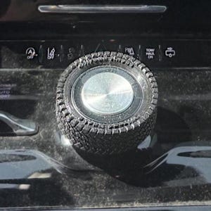 May include: Close-up of a black car control knob with a tire tread design. The knob is silver in the center and surrounded by a textured, black rubber-like material. The control panel has various symbols and text.