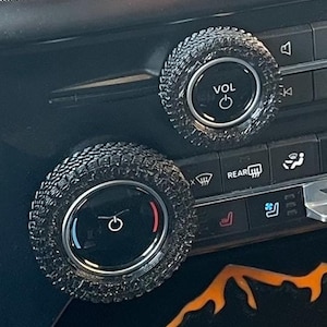 May include: Close-up of a car's dashboard with two round knobs. The knobs are black with a tire tread pattern and have a silver center. The knob on the left has a temperature gauge with red and blue markings. The knob on the right has the word "VOL" written on it.