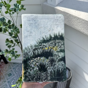 May include: A hand-painted Bible cover with a landscape design in green, white, and yellow. The cover features the words "Holy Bible" in a decorative font within a wreath.
