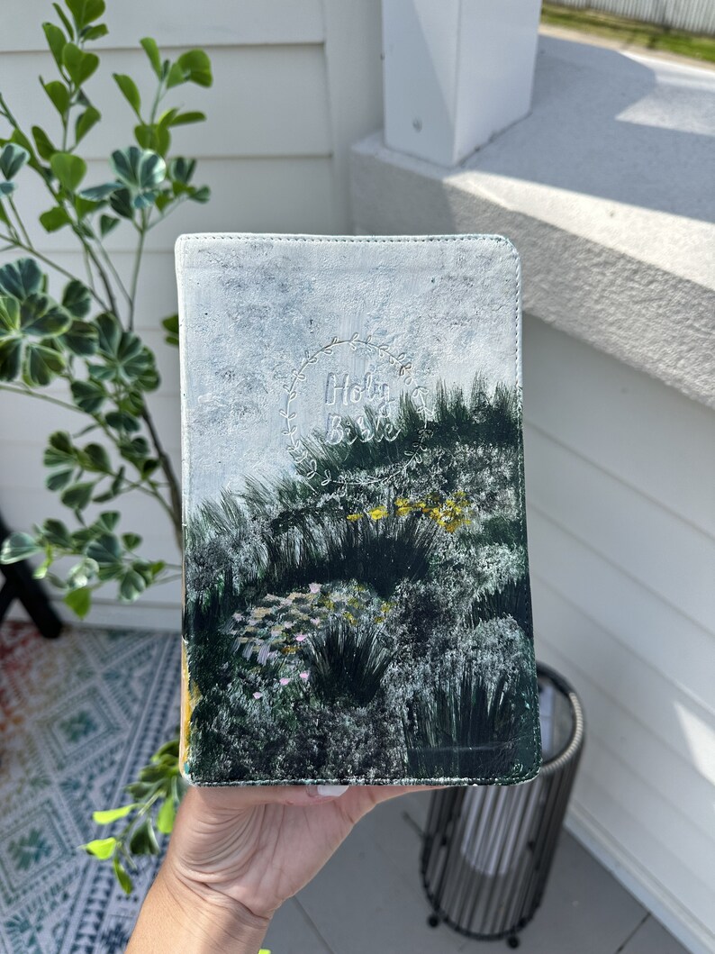 May include: A hand-painted Bible cover with a landscape design in green, white, and yellow. The cover features the words "Holy Bible" in a decorative font within a wreath.