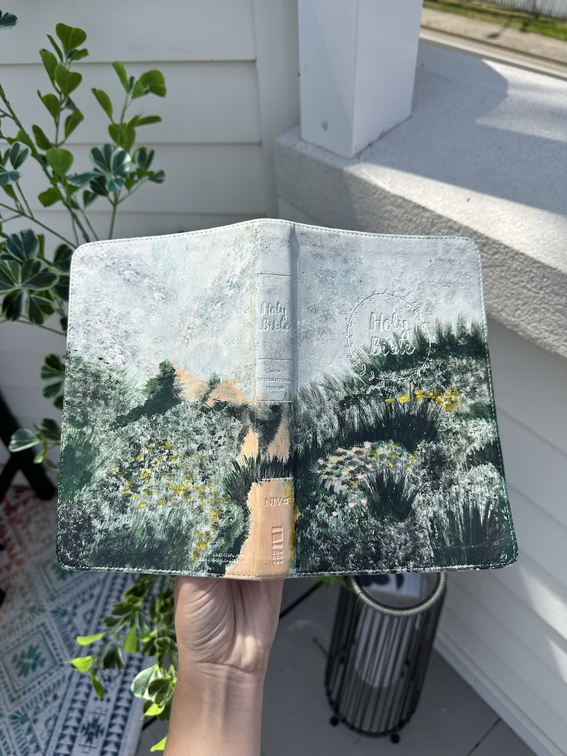 May include: A leather Bible cover with an original landscape painting. The cover features a path winding through green and yellow foliage, under a cloudy sky. The spine reads "Holy Bible" and "NIV".