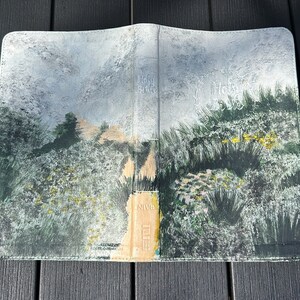 May include: A Bible cover featuring a landscape painting. The artwork shows a path through a field of green and yellow plants, under a cloudy sky. The words "Holy Bible" are visible on the cover.