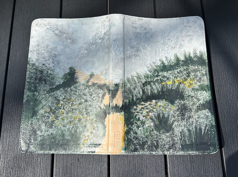 May include: A Bible cover featuring a landscape painting. The artwork shows a path through a field of green and yellow plants, under a cloudy sky. The words "Holy Bible" are visible on the cover.