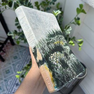 May include: A hardcover Holy Bible with a painted landscape design. The cover features a forest scene in shades of green, yellow, and beige. The spine is light blue with the words "Holy Bible" embossed.