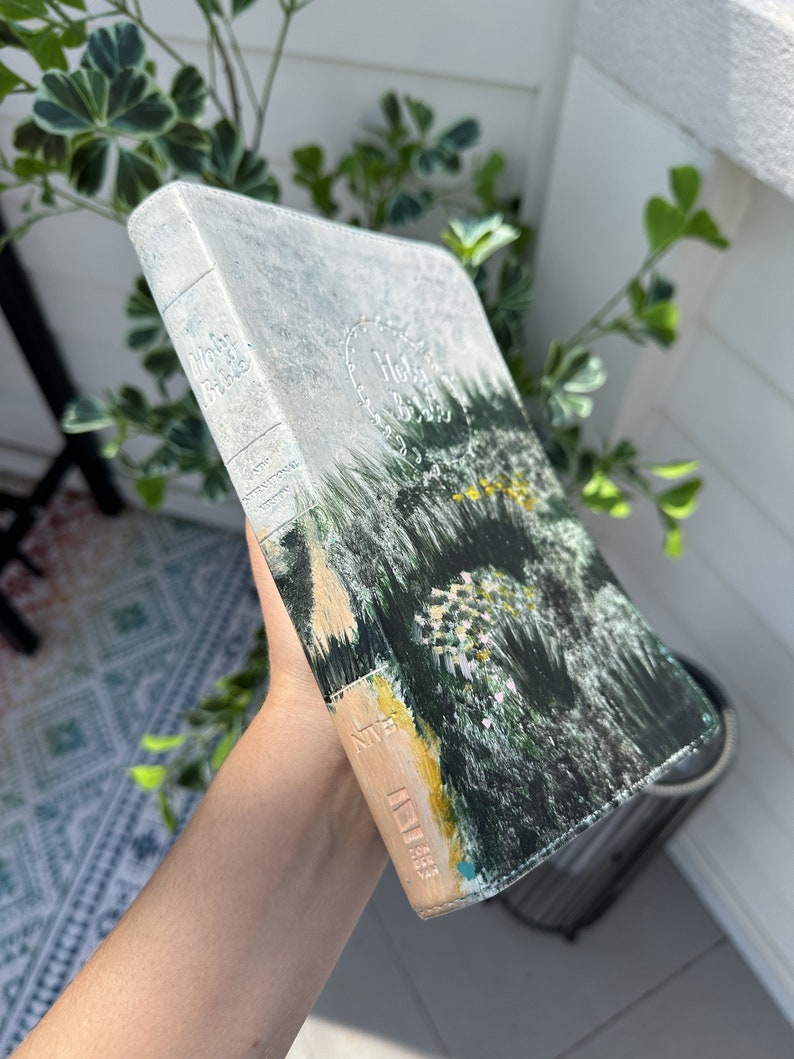May include: A hardcover Holy Bible with a painted landscape design. The cover features a forest scene in shades of green, yellow, and beige. The spine is light blue with the words "Holy Bible" embossed.