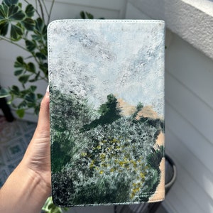 May include: A rectangular book cover featuring a landscape painting. The artwork shows a forest with green and yellow foliage, a tan hill, and a cloudy sky. The cover is held by a hand, and the edges are stitched.