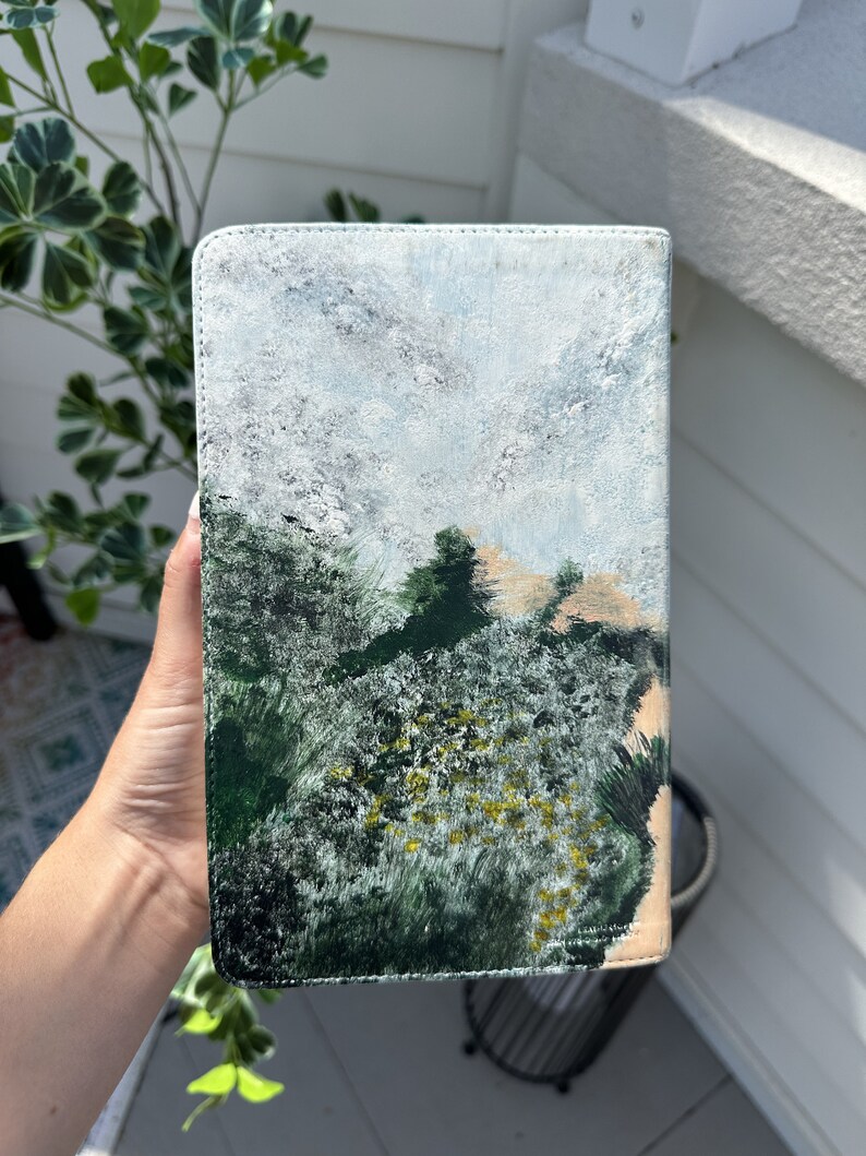 May include: A rectangular book cover featuring a landscape painting. The artwork shows a forest with green and yellow foliage, a tan hill, and a cloudy sky. The cover is held by a hand, and the edges are stitched.