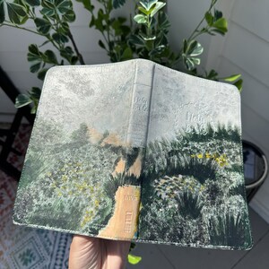 May include: An open Bible with a painted landscape design. The cover features a detailed illustration of a path winding through a field of green and yellow flowers. The spine is embossed with the words "Holy Bible".